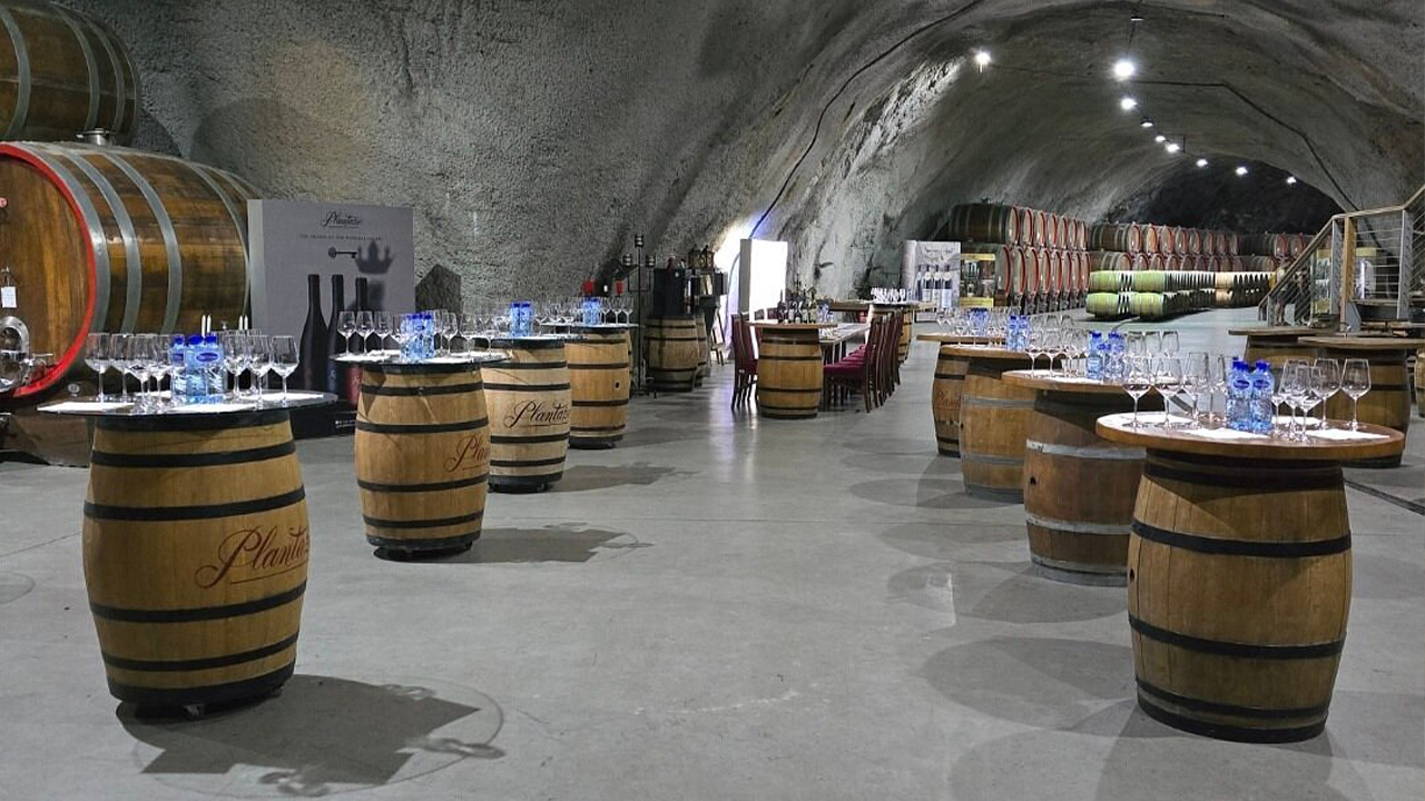 Barrel tables arranged for wine tasting inside a large underground hall filled with wine casks.